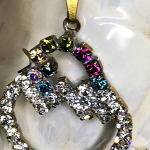 May include: A gold pendant with a heart-shaped design. The heart is outlined with clear rhinestones and has colorful rhinestones in the center. The colors of the rhinestones are green, blue, pink, and purple.