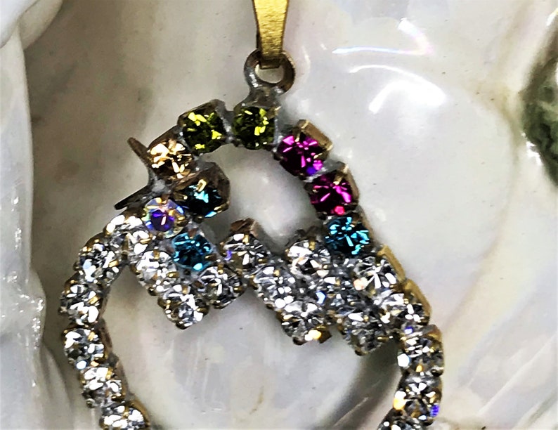 May include: A gold pendant with a heart-shaped design. The heart is outlined with clear rhinestones and has colorful rhinestones in the center. The colors of the rhinestones are green, blue, pink, and purple.