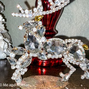 May include: A decorative reindeer figurine constructed from clear faceted crystals and rhinestones set in metal. The antlers, body, and legs are encrusted with various shapes of clear stones, with small yellow accents on the head and tail.