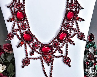 May include: A multi-strand bib necklace featuring deep red faceted rhinestones and large oval red cabochon stones set in a dark metal frame. The intricate design includes dangling elements and the text Me and Moma in the bottom left corner.
