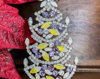 May include: A decorative Christmas tree ornament. The tree is made of silver-colored metal and adorned with clear, purple, and yellow gemstones. The tree is on a gold-colored base and has a star on top.