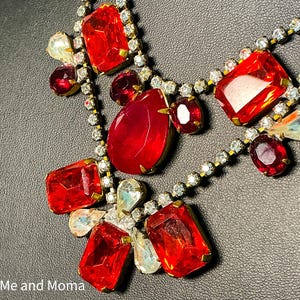 May include: A necklace featuring large, faceted red glass stones in rectangular, pear, and oval shapes, accented by smaller clear iridescent stones and clear rhinestones set in a gold-tone metal frame. The text Me and Moma is visible at the bottom.