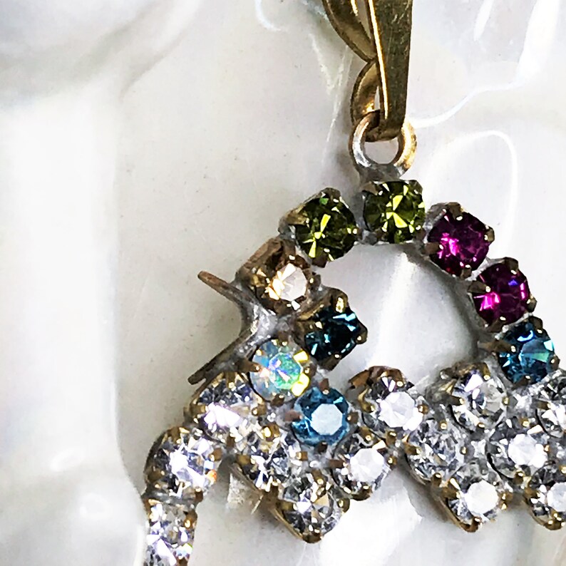 May include: A gold pendant with a heart-shaped design, featuring a rainbow of colorful rhinestones. The pendant is adorned with clear, green, blue, pink, and iridescent stones.