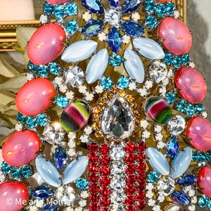 May include: Close-up of a vintage brooch with a complex design. The brooch features pink, blue, and clear gemstones, with a central teardrop-shaped crystal. The design includes oval and leaf-shaped stones, and red and white accents. The brooch has a gold-tone metal base.