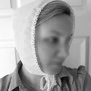 May include: A white cotton bonnet with lace trim, worn over a striped shirt. The bonnet has a bow tied at the back of the neck.