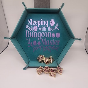 May include: A teal, hexagonal dice tray with the text "Sleeping with the Dungeon Master" in purple. Several gold and burgundy dice are inside the tray and next to it. The tray is made of a soft material and has snaps to keep it flat.