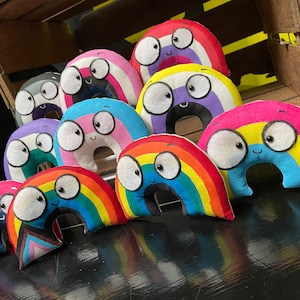 May include: A collection of rainbow-colored plush toys shaped like rainbows with smiling faces and large eyes. The toys are arranged in a row on a black surface.