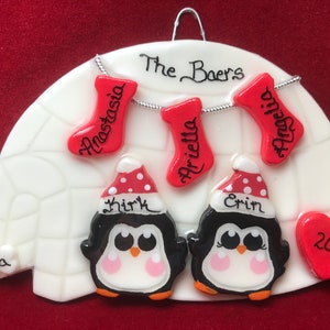May include: A personalized Christmas ornament featuring a white igloo shape with two penguin figures wearing Santa hats. Red stockings with names hang above. The ornament includes the family name "The Baers" and a red heart with the year 2019.