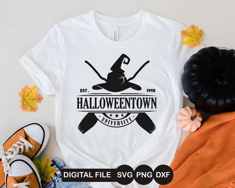 K&ouml;nnte beinhalten: Ein wei&szlig;es T-Shirt mit einem schwarzen Grafikdesign eines Hexenhutes mit gekreuzten Besen. Die Grafik enth&auml;lt auch den Text "EST. 1998 HALLOWEENTOWN UNIVERSITY".