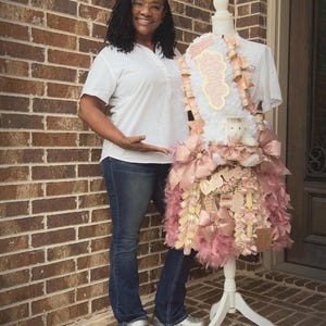 May include: A woman stands next to a decorative sash mum on a white stand. The mum is adorned with pink ribbons, feathers, and a plush bear. The text "THE Sash Mum Tutorial" is at the top of the image.
