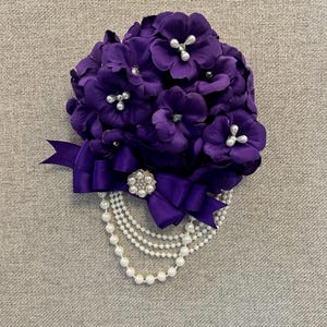 May include: A purple flower bouquet with a pearl necklace and a purple ribbon. The bouquet is made of fabric flowers and has a pearl brooch in the center.
