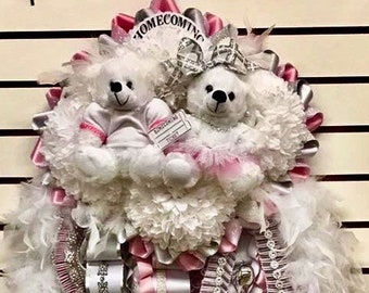 Puede incluir: Una mamá de regreso a casa con cintas blancas y rosadas, plumas blancas y dos osos de peluche blancos. Un oso de peluche lleva un tutú rosa y el otro lleva una camiseta de fútbol rosa y blanca. La mamá tiene un amuleto plateado con la palabra "Homecoming" en él.