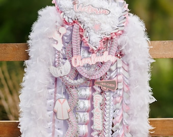 May include: A white and pink cheerleading graduation lei with a star-shaped Texas cutout, the name "Victoria" and the words "Cheerleader" and "Class of 2024". The lei also includes a white feather boa, a pink bow, and a white and pink ribbon with the words "All American Cheer".