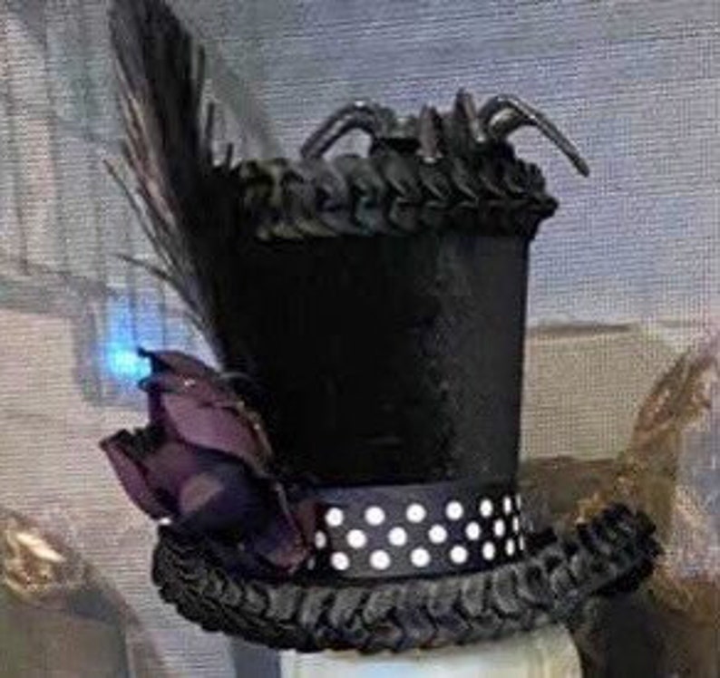 May include: A black top hat with a white polka dot band, a purple flower, and a black feather.  The hat is decorated with black spiders.