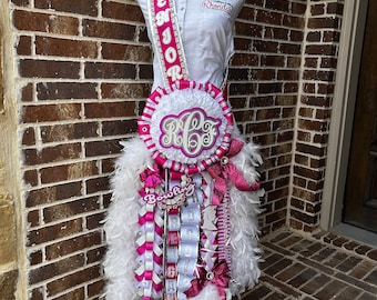 May include: A white and pink graduation sash with a white feather boa and a white and pink flower arrangement. The sash has the word "Senior" written on it in silver glitter. The flower arrangement has a white and pink bow and a white and pink ribbon with the word "Bowling" written on it. The sash is on a white mannequin with a white shirt that says "Rhonda" on it.