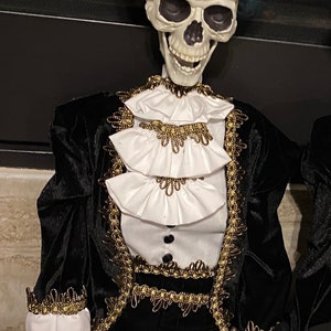 May include: A skeleton wearing a black velvet suit with gold trim and a black top hat with a purple flower and feather. The skeleton is smiling and has a white ruffled collar.