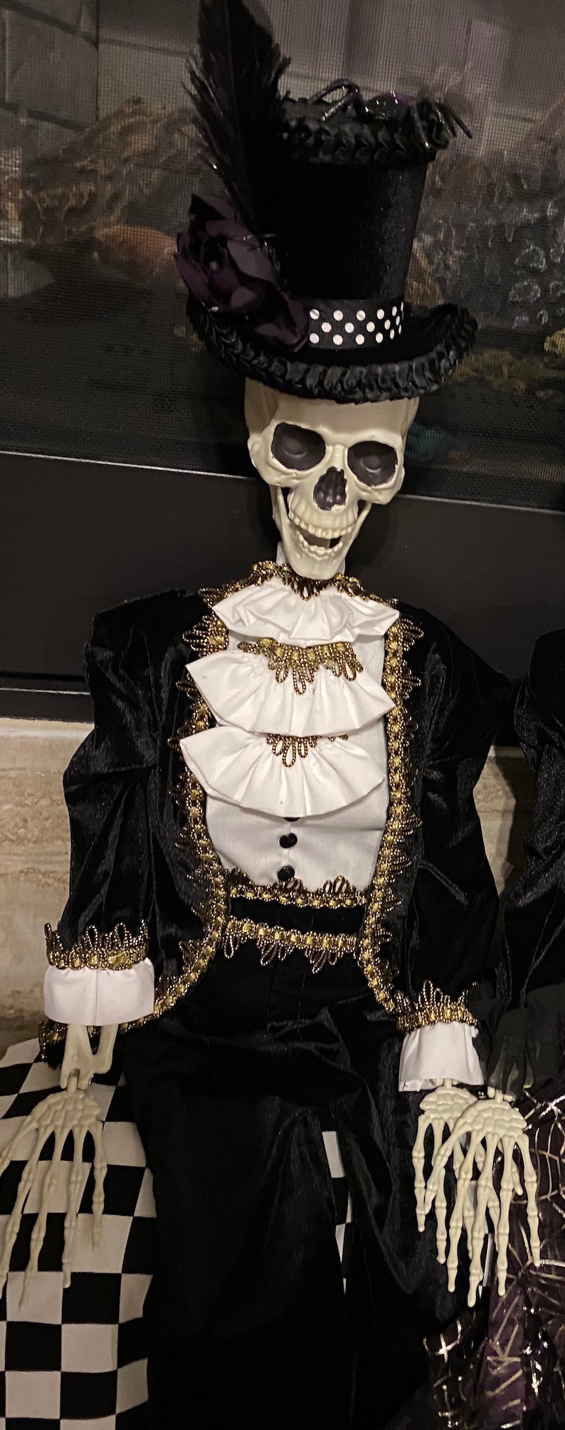 May include: A skeleton wearing a black velvet suit with gold trim and a black top hat with a purple flower and feather. The skeleton is smiling and has a white ruffled collar.