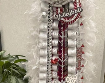 Puede incluir: Un lei de graduación blanco y rojo con un borde de plumas blancas. El lei tiene una flor blanca con un gorro de graduación negro en la parte superior. El lei también tiene una cinta roja y blanca con la palabra "Volleyball" en ella. El lei tiene una cinta blanca con el nombre "Zaria" en ella. El lei tiene una cinta blanca con el año "Senior 2023" en ella.