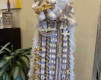 May include: A white and gold homecoming mum with a white teddy bear wearing a gold dress. The mum has white feathers, gold and white ribbons, and the words "Varsity Tennis" and "Senior" on it.