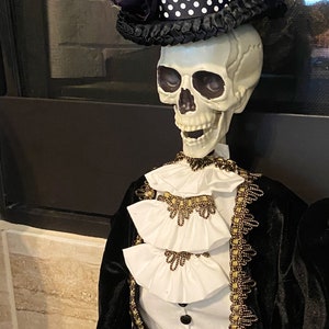 May include: A skeleton wearing a black velvet suit with gold trim and a white ruffled shirt. The skeleton is wearing a black top hat with a white polka dot band and a purple flower.
