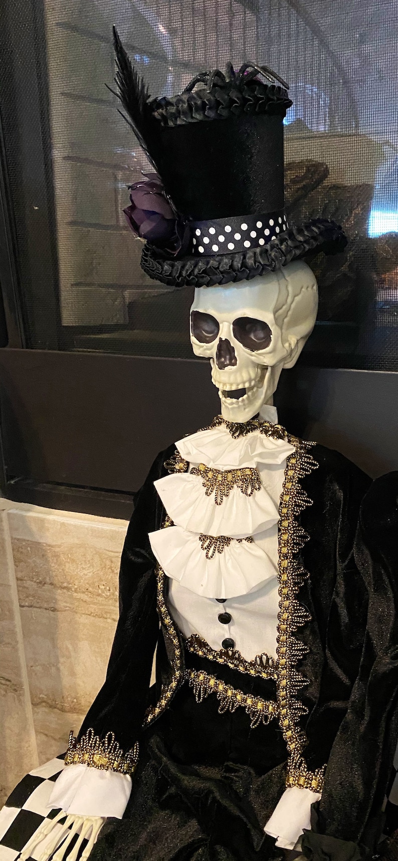 May include: A skeleton wearing a black velvet suit with gold trim and a white ruffled shirt. The skeleton is wearing a black top hat with a white polka dot band and a purple flower.