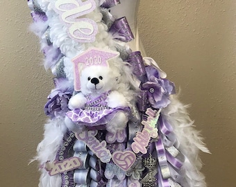 Puede incluir: Un collar de graduación blanco y morado con un oso de peluche blanco que lleva un vestido morado y un gorro de graduación. El collar tiene plumas blancas, cintas moradas y plateadas, y las palabras "Senior" y "2020".