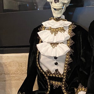 May include: A skeleton wearing a black velvet suit with gold trim and a white ruffled shirt. The skeleton is wearing a black top hat with a white polka dot band and a feather.