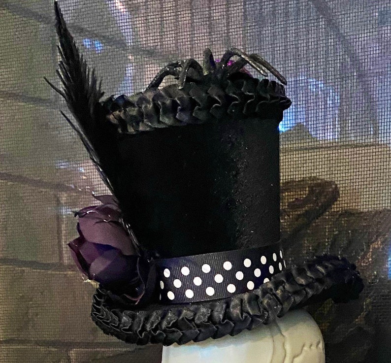 May include: A black top hat with a black feather, a purple flower, and a black and white polka dot ribbon. A black spider sits on top of the hat.