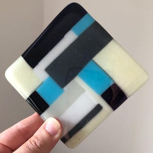 May include: Square glass coaster with a geometric design. The coaster features a pattern of black, white, light blue, and cream-colored rectangular shapes. The coaster is held in a hand.
