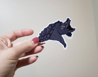 May include: A sticker featuring a dark gray snapping turtle with its mouth open. The turtle has a detailed, textured shell and is set against a white background. The sticker is held by a hand with painted nails.