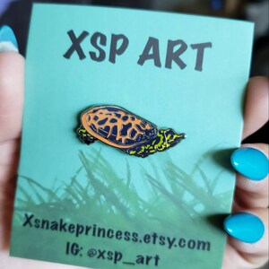 May include: A turtle enamel pin with a black outline, orange and black shell, and green accents. The pin is on a teal card with the text "XSP ART" and "Xsnakeprincess.etsy.com" and the Instagram handle "@xsp_art".