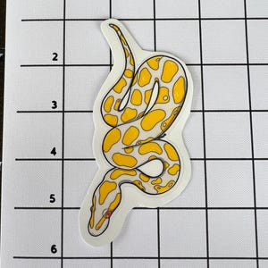 May include: A white sticker featuring a yellow and black snake design. The snake has a pattern of yellow spots and a red eye. The sticker is on a white grid background.