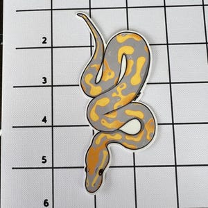 May include: A sticker featuring a stylized snake with a grey body and yellow and orange markings. The snake is coiled and has a white border. The sticker is approximately 10 cm long.