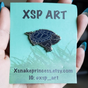 May include: A silver-colored turtle enamel pin with a dark blue shell, displayed on a light green card. The card has the text "XSP ART" at the top and "Xsnakeprincess.etsy.com" and "IG: @xsp_art" at the bottom. The pin is held in a hand.