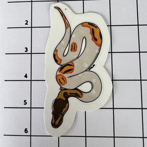 May include: A sticker depicting a stylized snake illustration. The snake has a light grey body with orange and brown markings. The snake's head is brown, and the sticker has a white border.