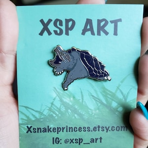 May include: A gray enamel pin featuring a snapping turtle with its mouth open. The pin is on a teal card with the text "XSP ART", "Xsnakeprincess.etsy.com", and "IG: @xsp_art".