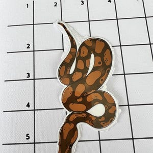 May include: A sticker depicting a brown and tan snake with a white border. The snake has a detailed pattern of dark brown spots on a lighter brown background. The sticker is a decorative item.
