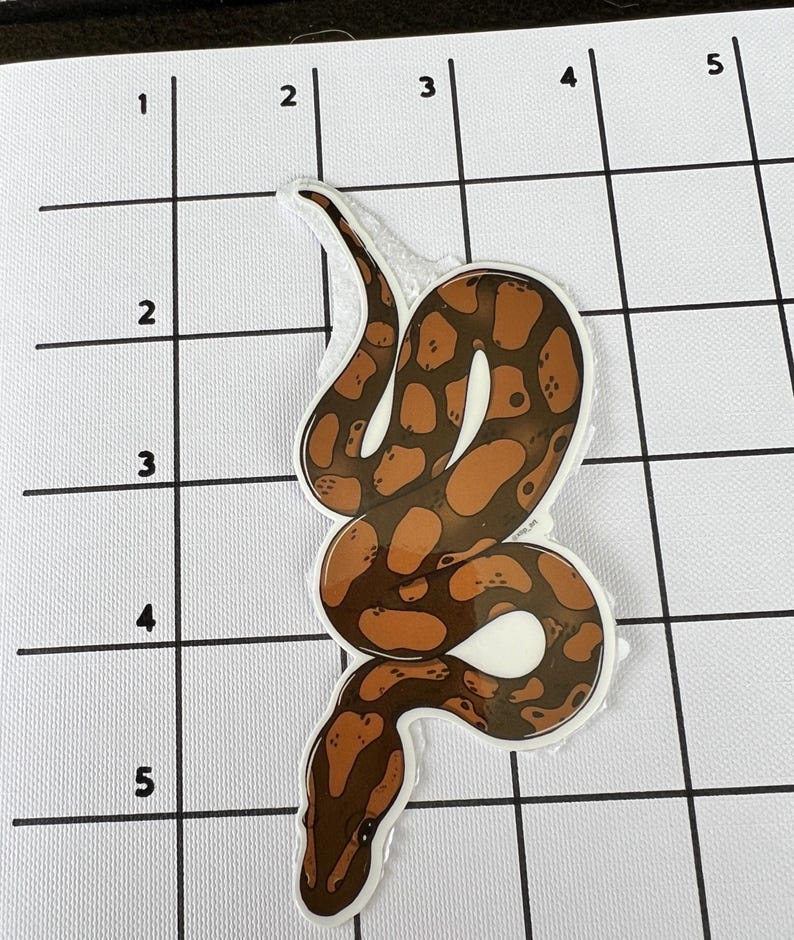 May include: A sticker depicting a brown and tan snake with a white border. The snake has a detailed pattern of dark brown spots on a lighter brown background. The sticker is a decorative item.