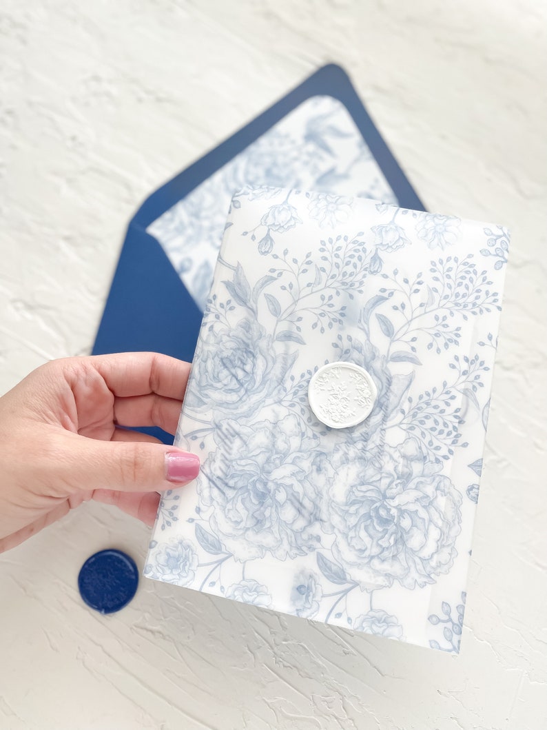 Puede incluir: Un sobre blanco con un patr&oacute;n floral azul y un sello de cera blanco. El sobre est&aacute; siendo sostenido por una mano.