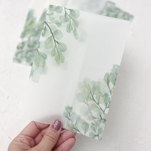 Puede incluir: Una hoja de papel transl&uacute;cido con un dise&ntilde;o de acuarela de hojas verdes. El papel est&aacute; doblado por la mitad y tiene un espacio en blanco en el centro.