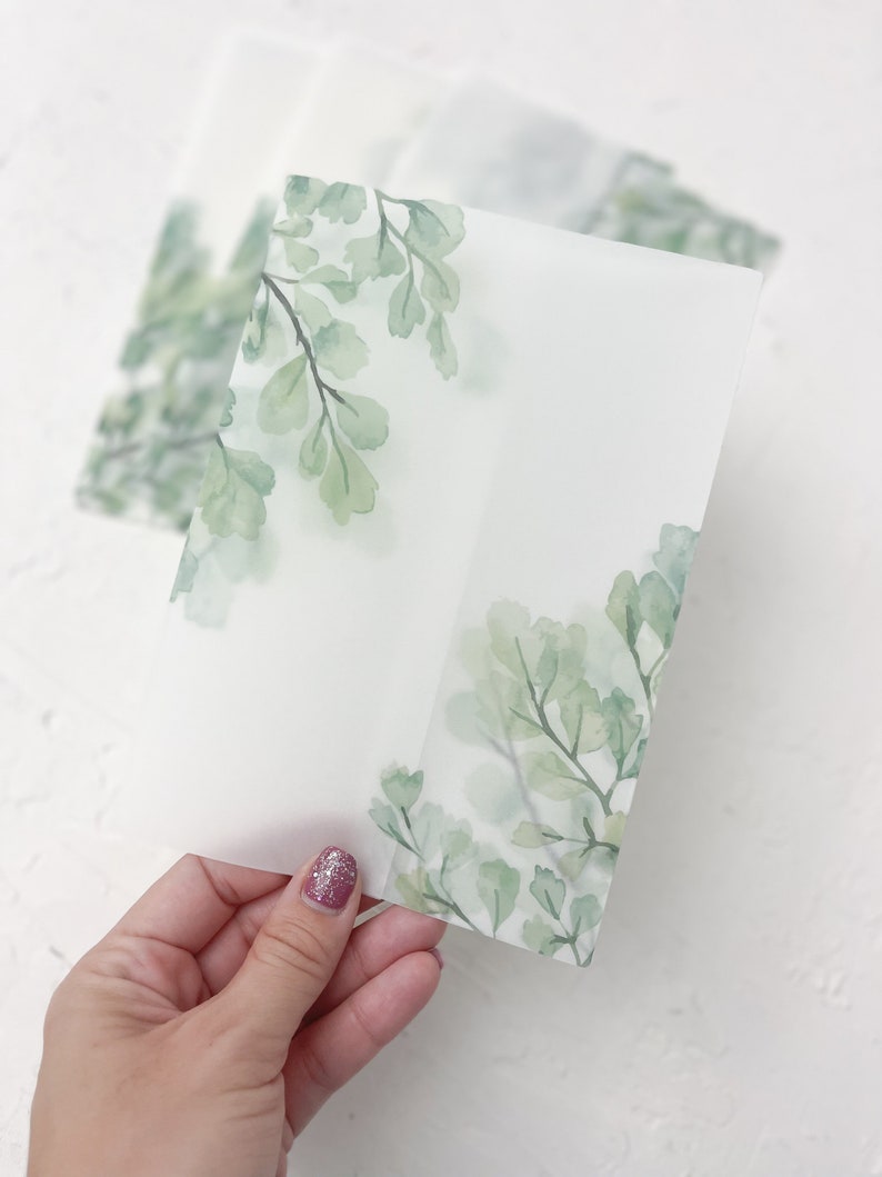Puede incluir: Una hoja de papel transl&uacute;cido con un dise&ntilde;o de acuarela de hojas verdes. El papel est&aacute; doblado por la mitad y tiene un espacio en blanco en el centro.