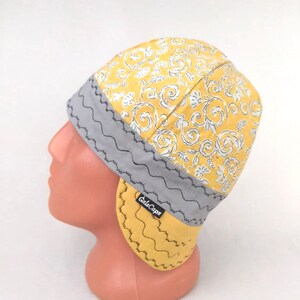 May include: A yellow and gray welding cap with a floral pattern and ear flaps. The cap has a gray band with a zig-zag stitch and the text "GalsCaps" is visible on the band.
