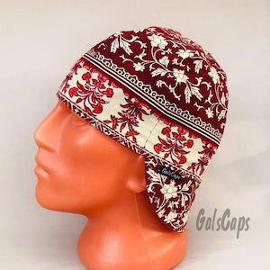May include: A burgundy welding cap with a floral pattern in white and red. The cap features a cream-colored band with a floral design and a decorative border. The brand name "GalsCaps" is visible on the cap.