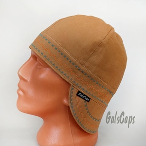 May include: A tan cap with ear flaps, adorned with blue and orange decorative stitching. The cap, displaying the text "GalsCaps", is presented on a mannequin head. The design includes a stitched border around the ear flaps.