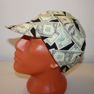 May include: A cap with a bill, featuring a repeating pattern of U.S. dollar bills in shades of green and black. The cap is made of fabric and is worn on a mannequin head.