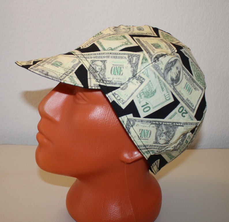 May include: A cap with a bill, featuring a repeating pattern of U.S. dollar bills in shades of green and black. The cap is made of fabric and is worn on a mannequin head.