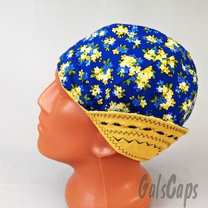 May include: A blue and yellow floral print welding cap. The cap features a vibrant blue background adorned with yellow and blue flower patterns. The brim is yellow with black stitching. The cap is designed to protect the head during welding.