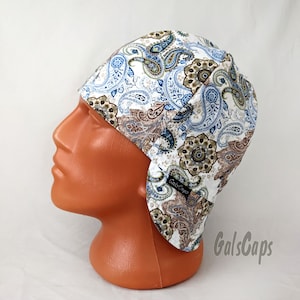 Op de afbeelding: Een witte lasmuts met een paisley-patroon in tinten blauw, bruin en groen. De muts heeft oorflappen en de tekst "GalsCaps" is aan de zijkant gedrukt.