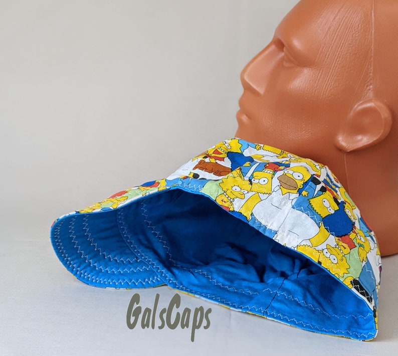 May include: A blue and yellow baseball cap with a cartoon print of characters from the Simpsons. The cap has a blue brim and a yellow crown.