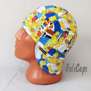 May include: A white welding cap with a blue and yellow pattern featuring characters from the Simpsons cartoon. The cap has a flap that can be worn down to protect the neck and ears. The brand name "GalsCaps" is printed on the flap.